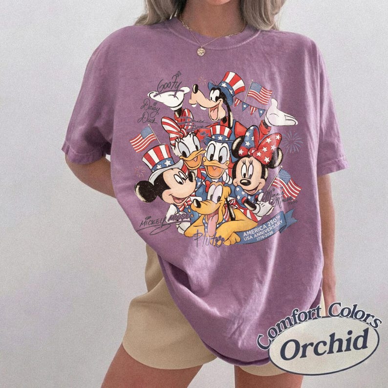 Puede incluir: Una camiseta morada Comfort Colors con un gr&aacute;fico de los personajes de Disney Mickey Mouse, Minnie Mouse, Donald Duck, Daisy Duck, Goofy y Pluto con atuendos patri&oacute;ticos. La camiseta tiene el texto "America 250th USA Anniversary 1776-2026" y "Comfort Colors Orchid".