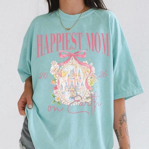 May include: A light blue t-shirt with the text "Happiest Mom on Earth" in pink. The shirt features a pink bow and a castle illustration surrounded by flowers. The numbers "20" and "26" are on either side of the castle.