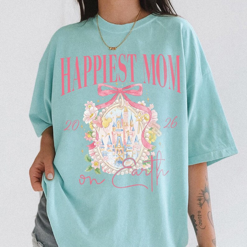 May include: A light blue t-shirt with the text "Happiest Mom on Earth" in pink. The shirt features a pink bow and a castle illustration surrounded by flowers. The numbers "20" and "26" are on either side of the castle.
