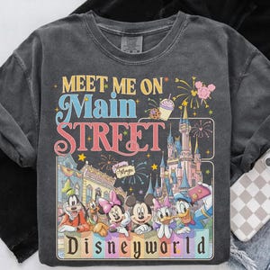 May include: Dark gray sweatshirt with a colorful graphic featuring Disney characters and the text "Meet Me On Main Street Disneyworld." The design includes a castle, fireworks, and various Disney characters.
