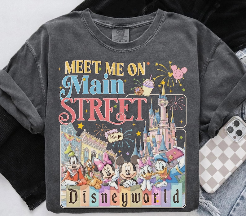 May include: Dark gray sweatshirt with a colorful graphic featuring Disney characters and the text "Meet Me On Main Street Disneyworld." The design includes a castle, fireworks, and various Disney characters.