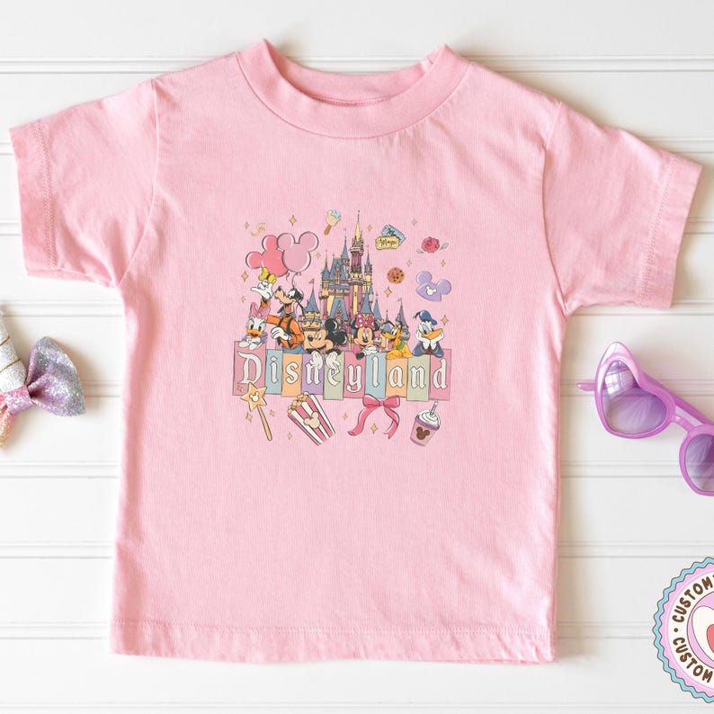 May include: A light pink t-shirt featuring a colorful graphic of Disneyland characters and a castle. The word "Disneyland" is printed in a rectangular box. Other graphics include a magic wand, popcorn, and a bow.