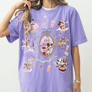 May include: A lavender t-shirt featuring a colorful graphic of Disney characters, including Mickey and Minnie Mouse, with the text "Best Day Ever." The design includes various Disney-themed elements.