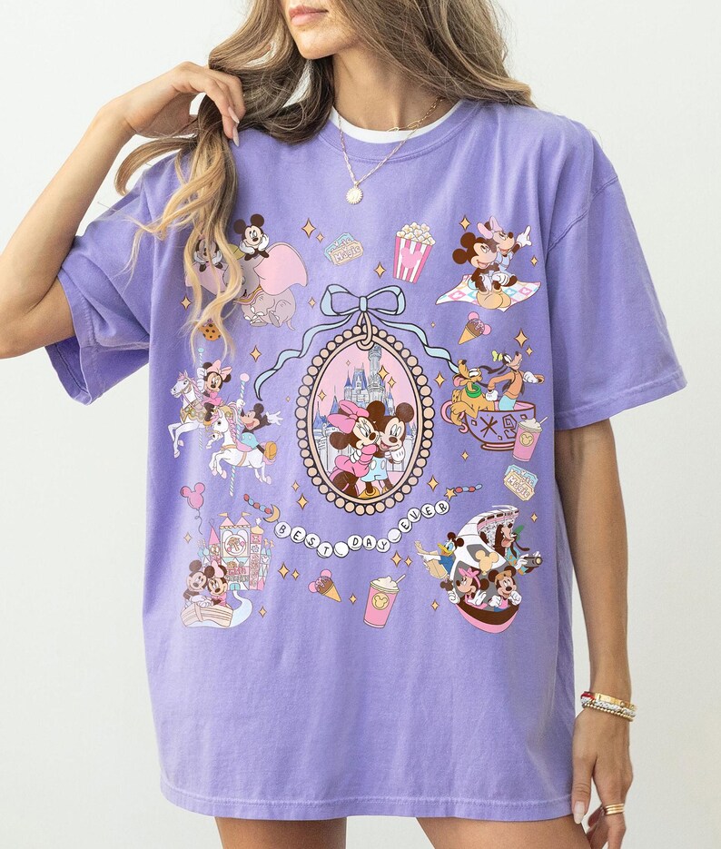 May include: A lavender t-shirt featuring a colorful graphic of Disney characters, including Mickey and Minnie Mouse, with the text "Best Day Ever." The design includes various Disney-themed elements.