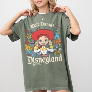 May include: Olive green t-shirt featuring a colorful Disneyland graphic with the text "Walt Disney's Disneyland Est 1955". The graphic includes a cartoon character with a red hat and various park attractions.