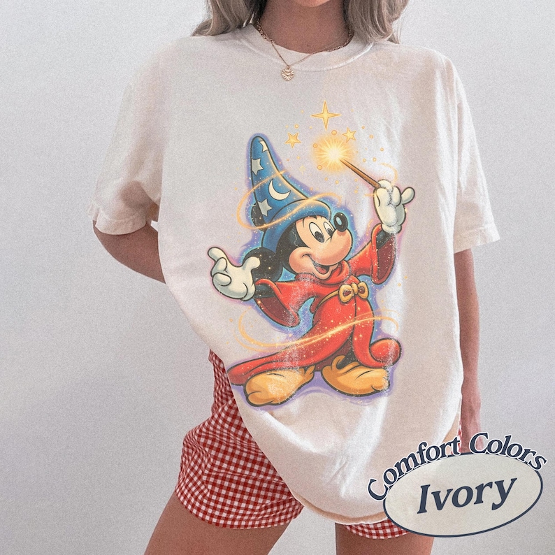 Pu&ograve; includere: T-shirt color avorio con una grafica di Topolino come mago, con un cappello blu e una veste rossa, che tiene in mano una bacchetta con un effetto a stella. La maglietta ha le parole "Comfort Colors" e "Ivory" stampate.