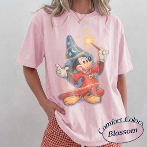 Pu&ograve; includere: T-shirt rosa chiaro Comfort Colors Blossom con un disegno di Topolino mago. Mickey indossa un cappello da mago blu con stelle, una veste rossa e tiene in mano una bacchetta magica con una stella luminosa. La maglietta ha un aspetto vintage.