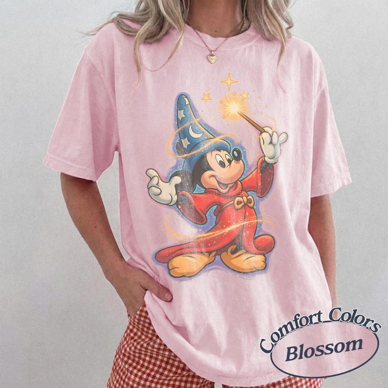 Pu&ograve; includere: T-shirt rosa chiaro Comfort Colors Blossom con un disegno di Topolino mago. Mickey indossa un cappello da mago blu con stelle, una veste rossa e tiene in mano una bacchetta magica con una stella luminosa. La maglietta ha un aspetto vintage.