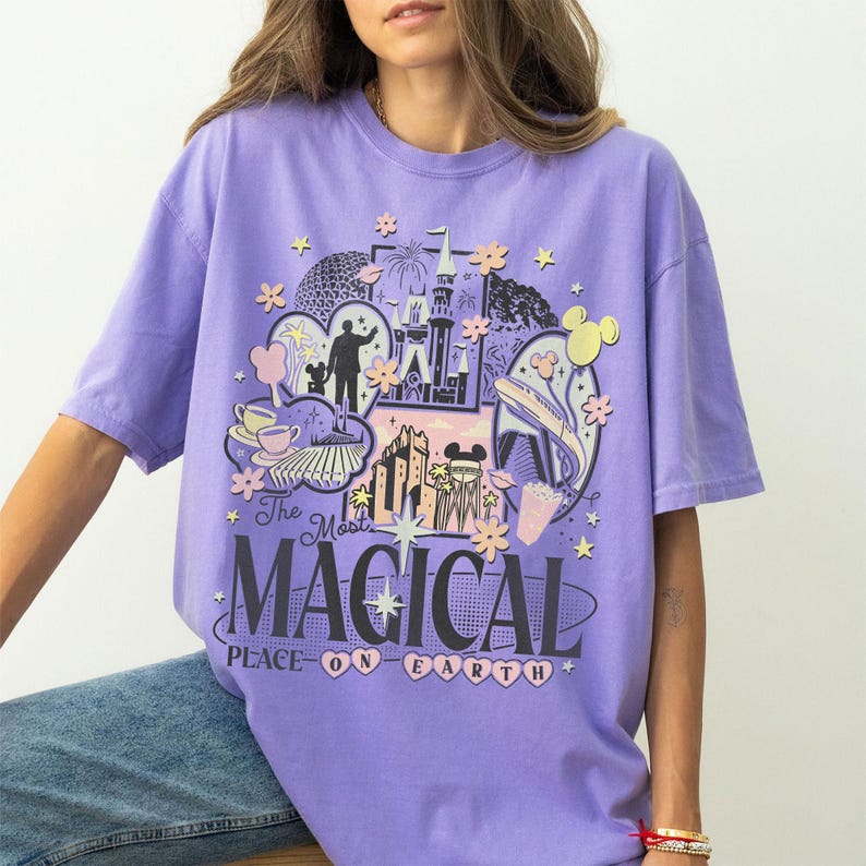 May include: A lilac t-shirt with a colourful graphic design. The design includes a silhouette of a person, a castle, and the text "The Most MAGICAL PLACE ON EARTH." The graphic incorporates floral and Mickey Mouse elements.