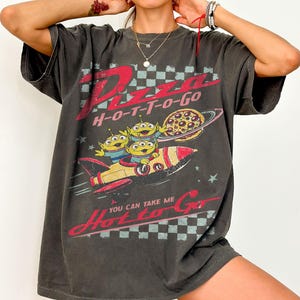 Pu&ograve; includere: T-shirt grigio scuro con grafica retr&ograve;. Il design presenta la scritta "Pizza H-O-T-T-O-Go", tre alieni verdi su un razzo e una pizza con un pianeta. La maglietta ha un aspetto vintage e usurato.