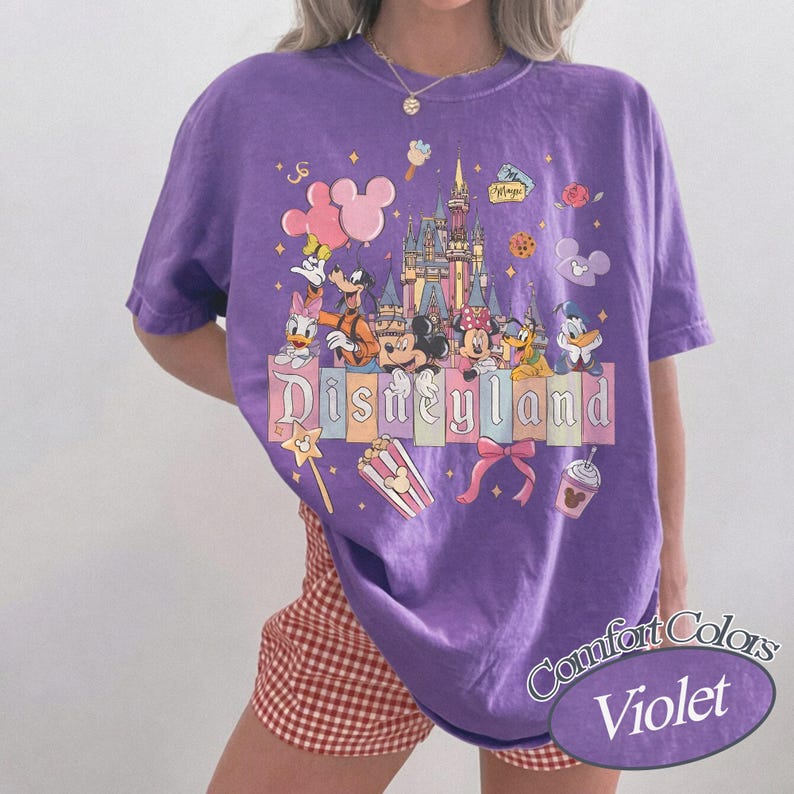 May include: A purple t-shirt with a Disneyland graphic featuring cartoon characters and a castle. The shirt has the words "Disneyland" and "Comfort Colors Violet" printed on it. The shirt is a crew neck.