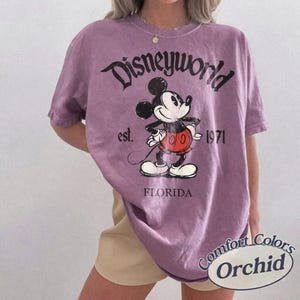 May include: A purple t-shirt with a Disneyworld graphic featuring Mickey Mouse. The design includes the text "Disneyworld," "est. 1971," and "Florida." The shirt is a Comfort Colors brand in the color Orchid.