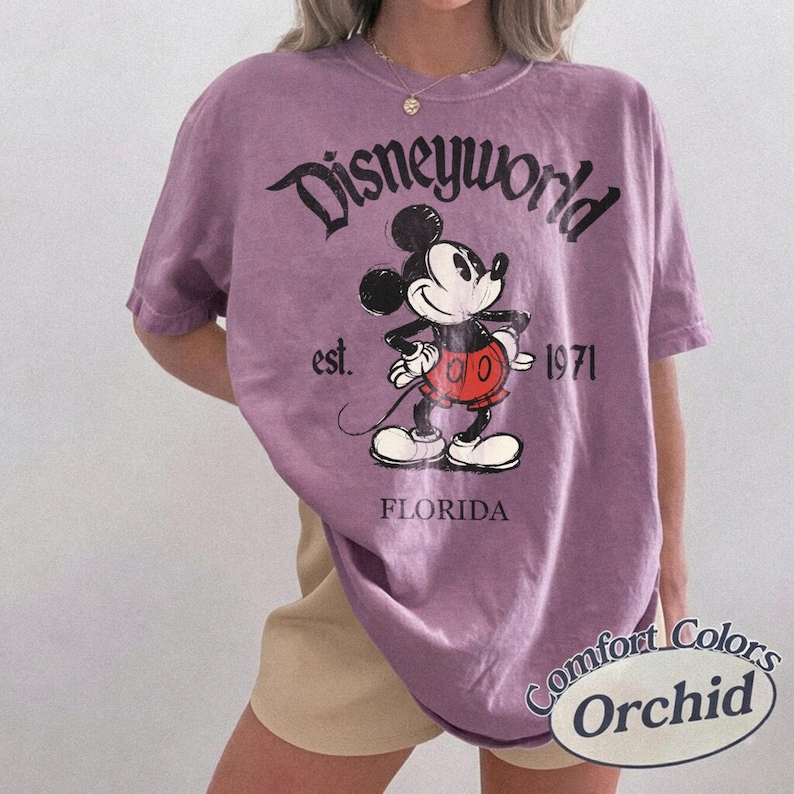 May include: A purple t-shirt with a Disneyworld graphic featuring Mickey Mouse. The design includes the text "Disneyworld," "est. 1971," and "Florida." The shirt is a Comfort Colors brand in the color Orchid.