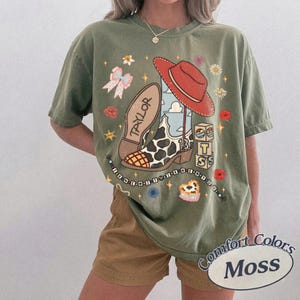 K&ouml;nnte beinhalten: Ein olivgr&uuml;nes T-Shirt mit einem Grafikdruck, der einen Cowboystiefel mit Kuhfleckenmuster und dem Namen Taylor, einen roten Cowboyhut, eine Perlenkette, kleine Bl&ouml;cke mit den Buchstaben T S sowie verschiedene Blumen- und Sternmotive zeigt.