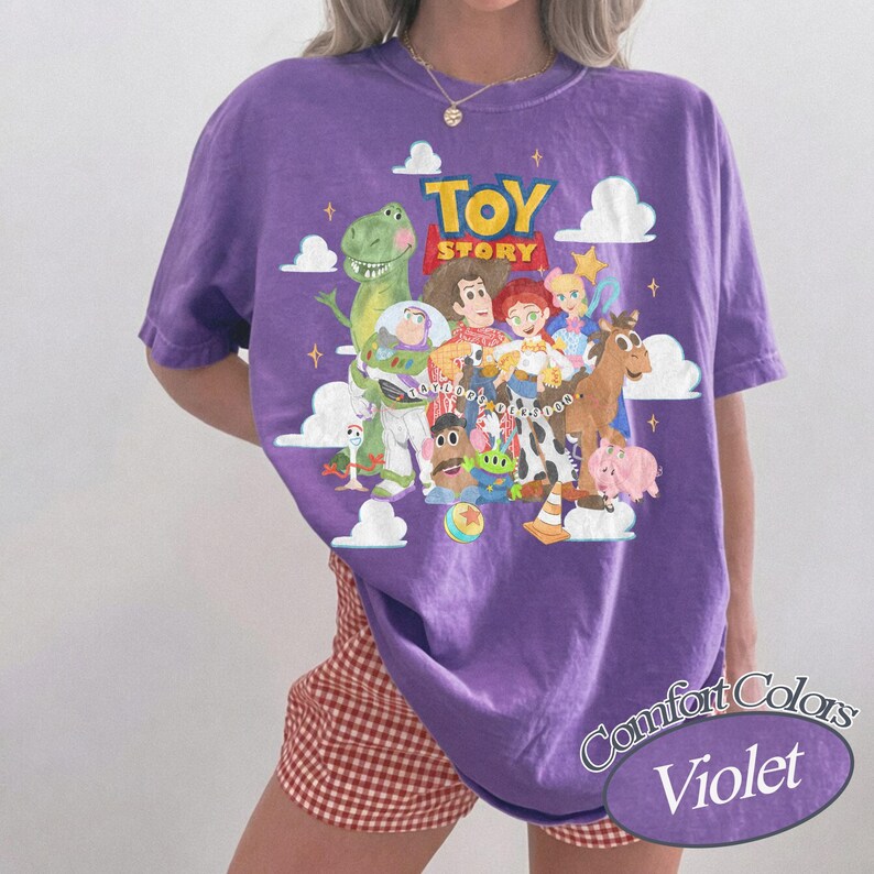 May include: A violet short-sleeve t-shirt featuring a colorful graphic print of Toy Story characters including Woody, Buzz Lightyear, Rex, and Jessie. The design includes white clouds, yellow stars, and the Toy Story logo. The shirt has a crew neckline.