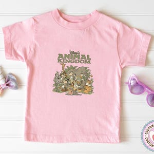 Puede incluir: Una camiseta rosa claro con un gr&aacute;fico de varios personajes y animales de Disney, incluyendo Mickey Mouse, una jirafa y un tigre. El texto "Disney's Animal Kingdom" est&aacute; impreso encima de los personajes en verde.