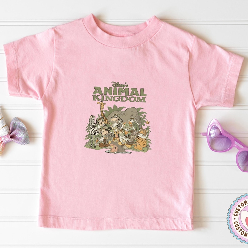 Puede incluir: Una camiseta rosa claro con un gr&aacute;fico de varios personajes y animales de Disney, incluyendo Mickey Mouse, una jirafa y un tigre. El texto "Disney's Animal Kingdom" est&aacute; impreso encima de los personajes en verde.
