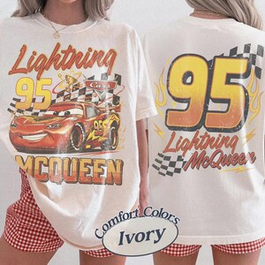 Puede incluir: Camiseta color marfil con un dise&ntilde;o de Rayo McQueen de la pel&iacute;cula Cars. La parte delantera muestra el coche con el texto "Lightning 95 McQueen". La parte trasera muestra "95 Lightning McQueen" con llamas y un dise&ntilde;o de bandera a cuadros.