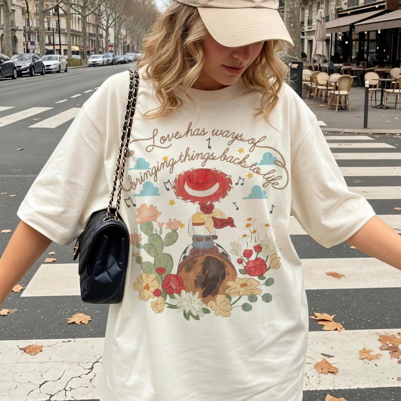 May include: An off-white t-shirt featuring a graphic of a cowgirl on a horse surrounded by cacti and flowers. The text reads, Love has ways of bringing things back to life. The shirt is paired with a black quilted shoulder bag with a gold chain strap.