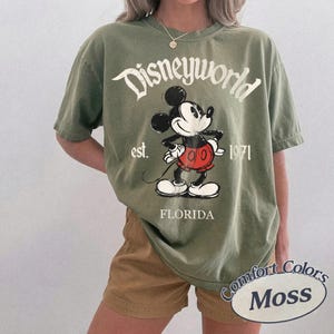 May include: Olive green t-shirt with a graphic of Mickey Mouse and the text "Disneyworld est. 1971 Florida." The shirt is a Comfort Colors Moss brand.