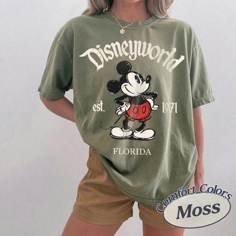 May include: Olive green t-shirt with a graphic of Mickey Mouse and the text "Disneyworld est. 1971 Florida." The shirt is a Comfort Colors Moss brand.