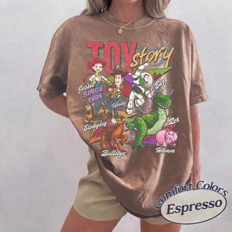 May include: A brown short-sleeve t-shirt featuring a vintage-style graphic of Toy Story characters including Jessie, Woody, Buzz Lightyear, Slinky Dog, Bullseye, Rex, and Hamm. The text reads Toy Story, Since 1995, and the names of each character.