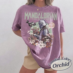 Może przedstawiać: Fioletowa koszulka z nadrukiem The Mandalorian i Grogu otoczonych kwiatami. Tekst "THE MANDALORIAN" i "THIS IS THE WAY" jest wydrukowany nad grafiką. Koszulka jest oznaczona jako "Comfort Colors Orchid".