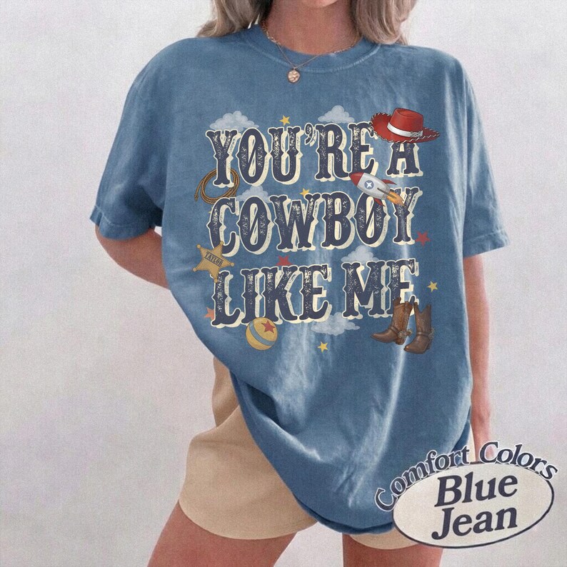 May include: A blue short-sleeve t-shirt featuring the text "YOU'RE A COWBOY LIKE ME" in a bold, distressed font. The graphic includes illustrations of a red cowboy hat, a rocket, a sheriff star, a ball, and brown cowboy boots with clouds.
