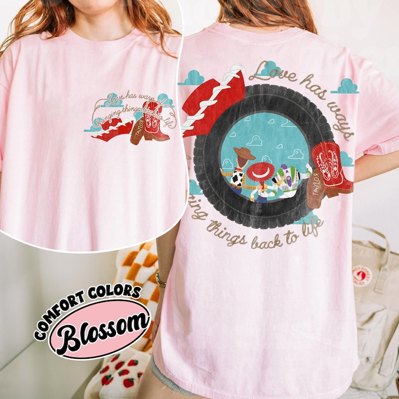 May include: A light pink t-shirt featuring a graphic of a black tyre framing cartoon characters, including a cowboy and a space ranger. The text "Love has ways bringing things back to life" is printed in brown script around the tyre and on the chest.