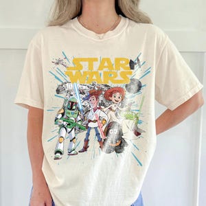 Puede incluir: Camiseta de color crema con un dise&ntilde;o gr&aacute;fico con las palabras "STAR WARS" en amarillo. El dise&ntilde;o incluye personajes de dibujos animados de las franquicias Star Wars y Toy Story, con detalles en azul y amarillo.