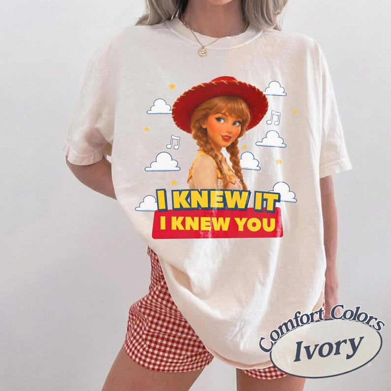 May include: An ivory short-sleeve t-shirt featuring a graphic of a character with braided hair wearing a red cowboy hat. The design includes white clouds, yellow stars, musical notes, and the text I KNEW IT I KNEW YOU in yellow and red.