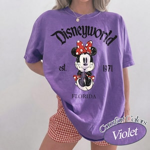 May include: A violet-colored t-shirt featuring a Minnie Mouse graphic with a red bow. The shirt has the text "Disneyworld" above the graphic, with "est. 1971" and "FLORIDA" below. The shirt is from the Comfort Colors brand.
