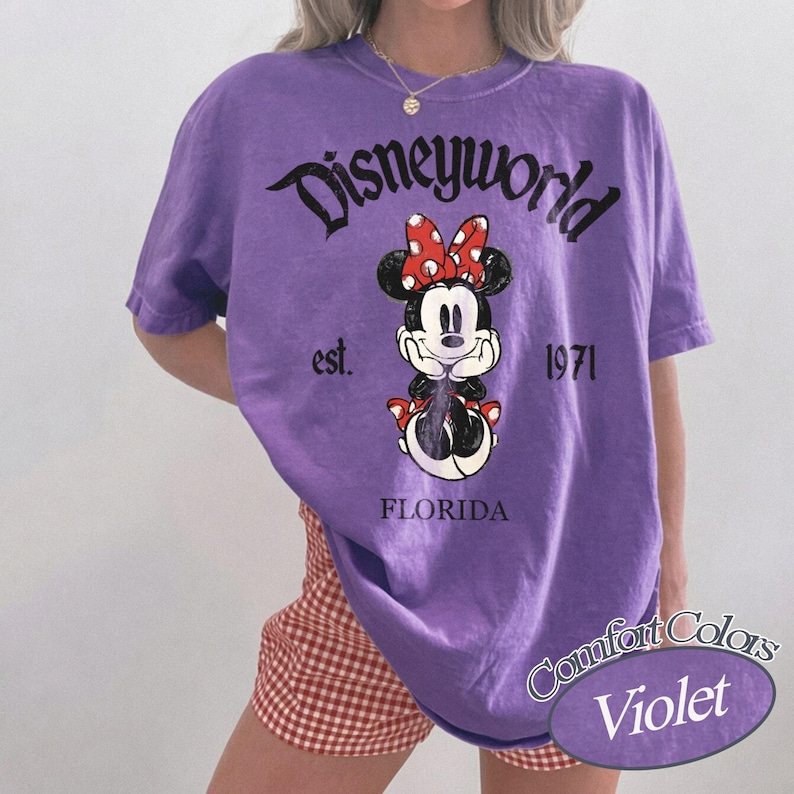 May include: A violet-colored t-shirt featuring a Minnie Mouse graphic with a red bow. The shirt has the text "Disneyworld" above the graphic, with "est. 1971" and "FLORIDA" below. The shirt is from the Comfort Colors brand.