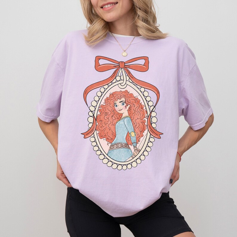 May include: A light purple t-shirt featuring a cartoon illustration of a character with red curly hair, wearing a blue dress, framed by a pearl-lined oval and a red bow. The shirt is a solid color.