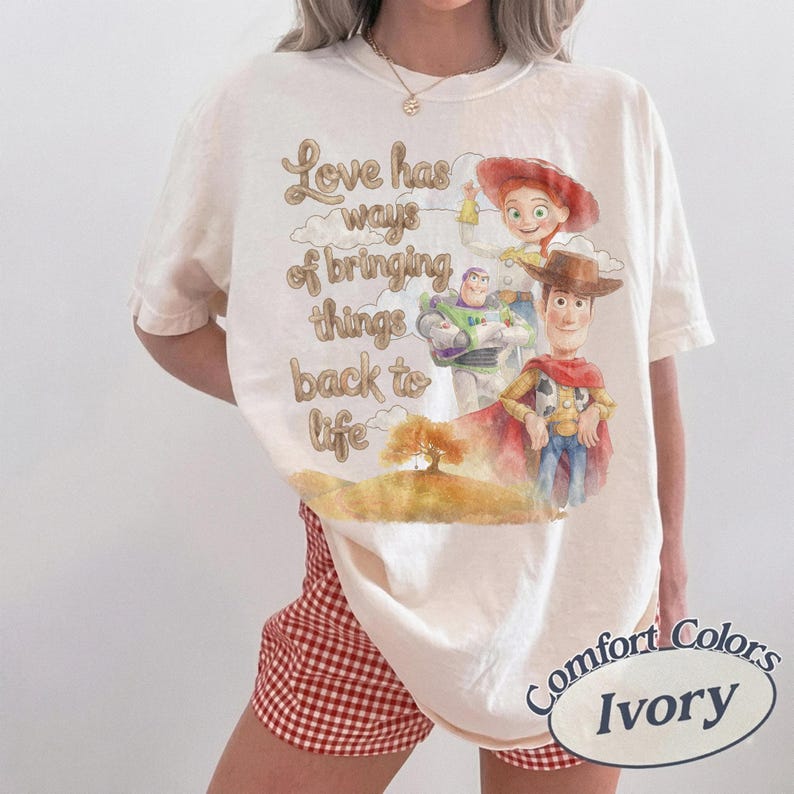 K&ouml;nnte beinhalten: Ein elfenbeinfarbenes Kurzarm-T-Shirt mit einem Grafikdruck von Woody, Buzz Lightyear und Jessie aus Toy Story. Der Text lautet, Love has ways of bringing things back to life, begleitet von einer Aquarell-Illustration eines Baumes.