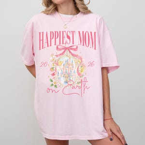 May include: A light pink t-shirt with the words "Happiest Mom" in a bold, pink font. Below the text is a pink bow and an oval design with a castle illustration. The words "20" and "26" flank the design, with "on Earth" below.