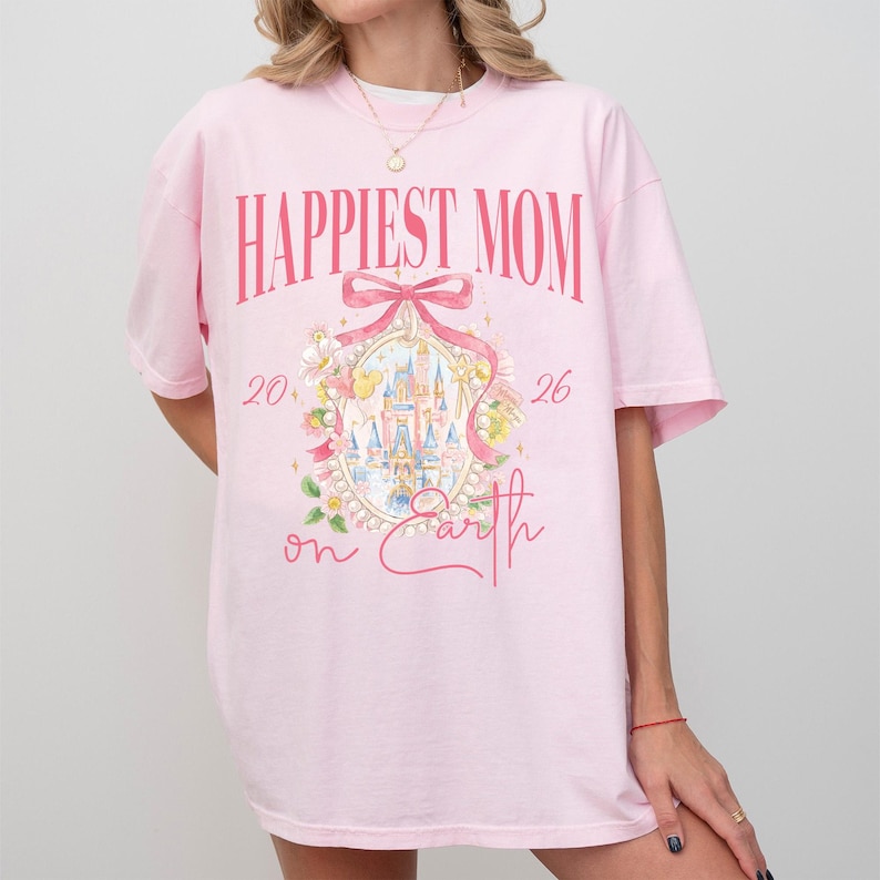 May include: A light pink t-shirt with the words "Happiest Mom" in a bold, pink font. Below the text is a pink bow and an oval design with a castle illustration. The words "20" and "26" flank the design, with "on Earth" below.
