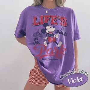 May include: A violet Comfort Colors t-shirt with a graphic of Mickey Mouse walking in front of a castle. The text reads "LIFE'S A WALK IN THE PARK" in red and white, with "Walt Disney World" below. The shirt is a Comfort Colors brand.
