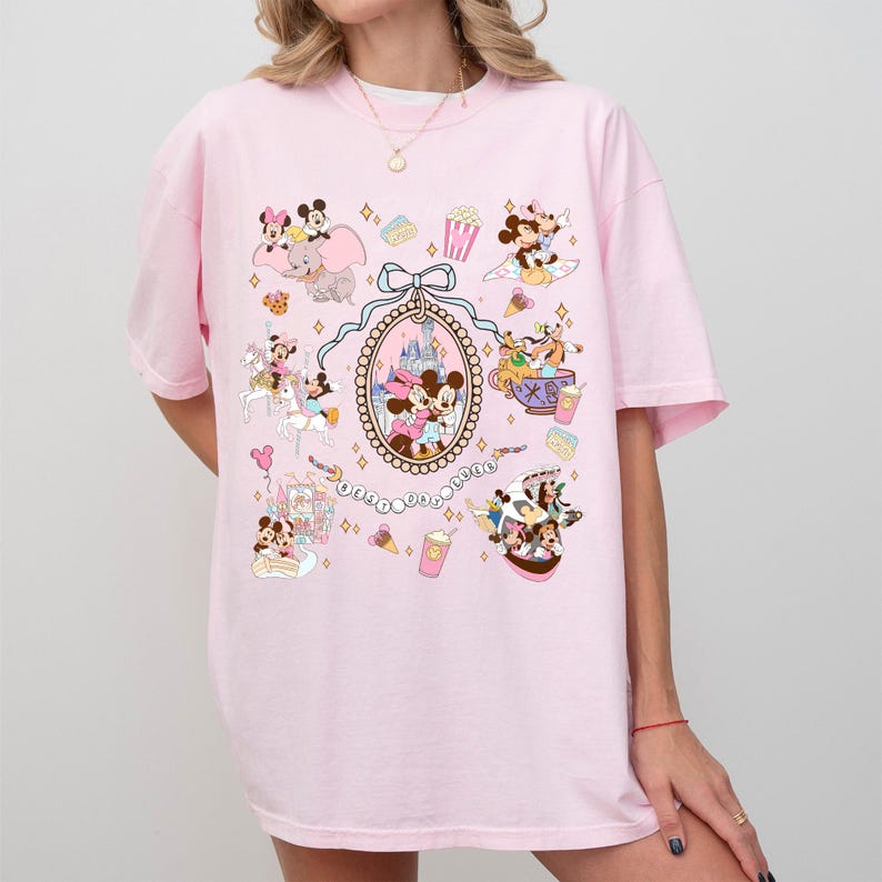 May include: A light pink t-shirt featuring a colorful graphic of Disney characters and park icons. The design includes Mickey and Minnie Mouse, Dumbo, and various park attractions. The shirt has short sleeves and a crew neck.
