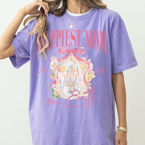 May include: A lilac-colored t-shirt with the words "Happiest Mom on Earth" in pink. The shirt features a graphic of a castle, flowers, and a bow. The text "2026" is also visible on the shirt.