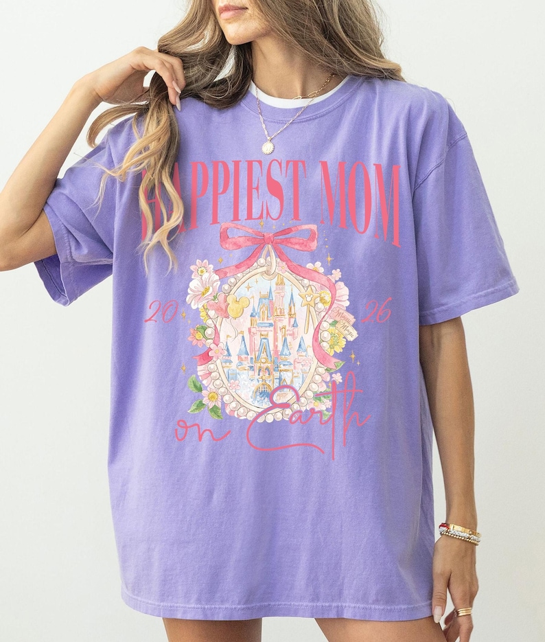 May include: A lilac-colored t-shirt with the words "Happiest Mom on Earth" in pink. The shirt features a graphic of a castle, flowers, and a bow. The text "2026" is also visible on the shirt.