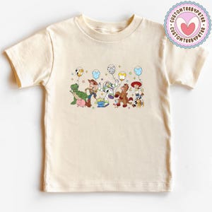 May include: Cream-colored t-shirt featuring a colorful graphic of Toy Story characters holding balloons. The design includes Woody, Buzz Lightyear, Rex, and other characters. The shirt has short sleeves and a crew neck.