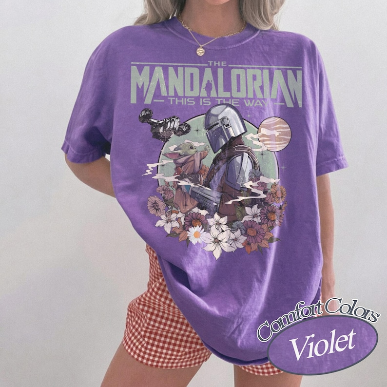 Może przedstawiać: Fioletowa koszulka Comfort Colors z grafiką The Mandalorian i Grogu otoczonych kwiatami. Tekst "THE MANDALORIAN" i "THIS IS THE WAY" jest wydrukowany nad obrazem. Koszulka jest w jednolitym fioletowym kolorze.