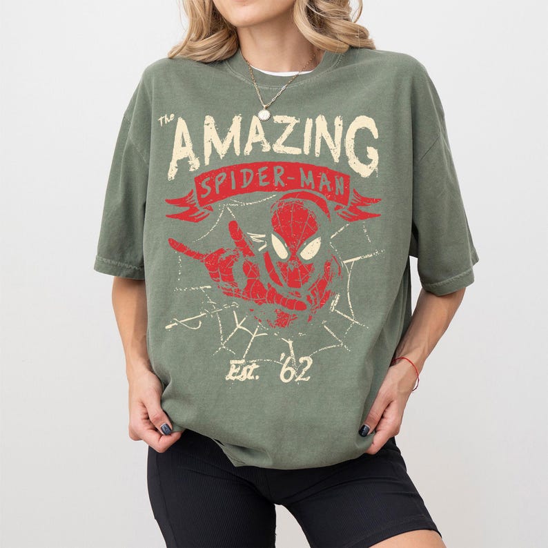 Pode incluir: Camiseta verde-oliva com um gr&aacute;fico desgastado do Homem-Aranha. O design inclui as palavras "The Amazing Spider-Man" em uma faixa, uma imagem vermelha do Homem-Aranha e "Est. '62". A camiseta tem mangas curtas e um corte folgado.