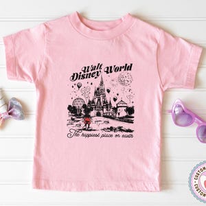 Peut inclure: Un t-shirt rose clair avec un dessin en noir et blanc du ch&acirc;teau de Cendrillon, des ballons et des feux d'artifice, avec le texte "Walt Disney World" et "The happiest place on earth".