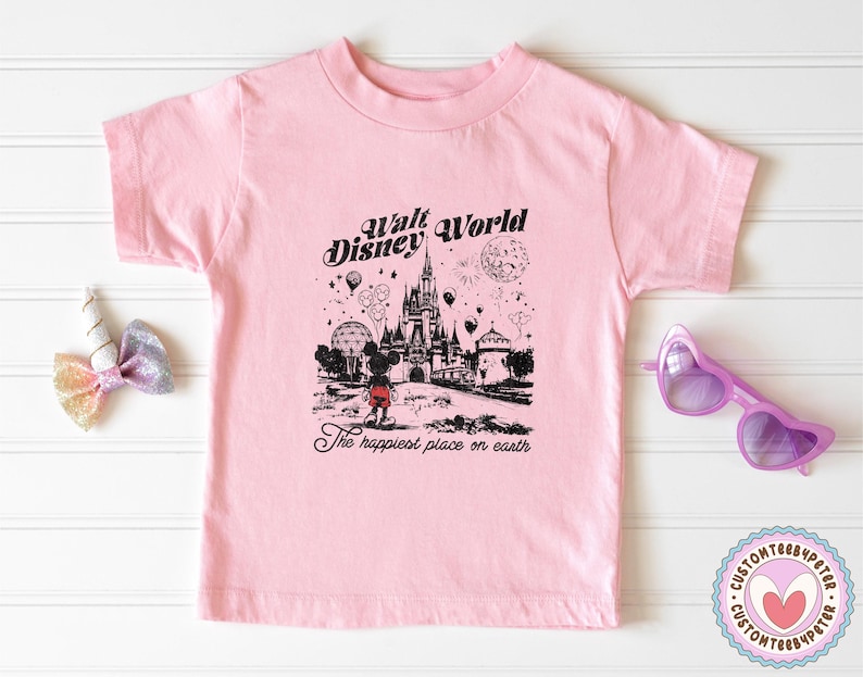 Peut inclure: Un t-shirt rose clair avec un dessin en noir et blanc du ch&acirc;teau de Cendrillon, des ballons et des feux d'artifice, avec le texte "Walt Disney World" et "The happiest place on earth".
