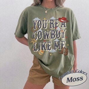 May include: An olive green short-sleeve t-shirt featuring the text "YOU'RE A COWBOY LIKE ME" in a dark blue font. The graphic includes a red cowboy hat, a rocket ship, a sheriff star, a ball, and brown cowboy boots, all set against a cloudy sky.