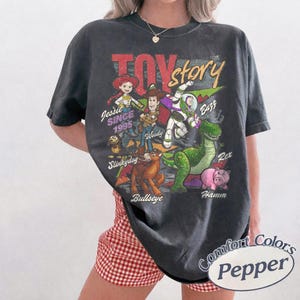 May include: A charcoal gray short-sleeve t-shirt featuring a vintage-style graphic of Toy Story characters including Jessie, Woody, Buzz Lightyear, Slinky Dog, Bullseye, Rex, and Hamm. The text reads Toy Story, Jessie, Since 1995, Woody, Buzz, Slinky Dog, Bullseye, Rex, and Hamm.