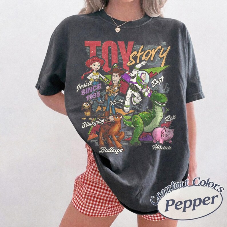May include: A charcoal gray short-sleeve t-shirt featuring a vintage-style graphic of Toy Story characters including Jessie, Woody, Buzz Lightyear, Slinky Dog, Bullseye, Rex, and Hamm. The text reads Toy Story, Jessie, Since 1995, Woody, Buzz, Slinky Dog, Bullseye, Rex, and Hamm.