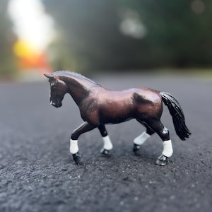 May include: A brown toy horse with a black mane and tail, and white leg wraps. The horse is posed in a walking stance on a dark surface, with a blurred background of green trees and sunlight.